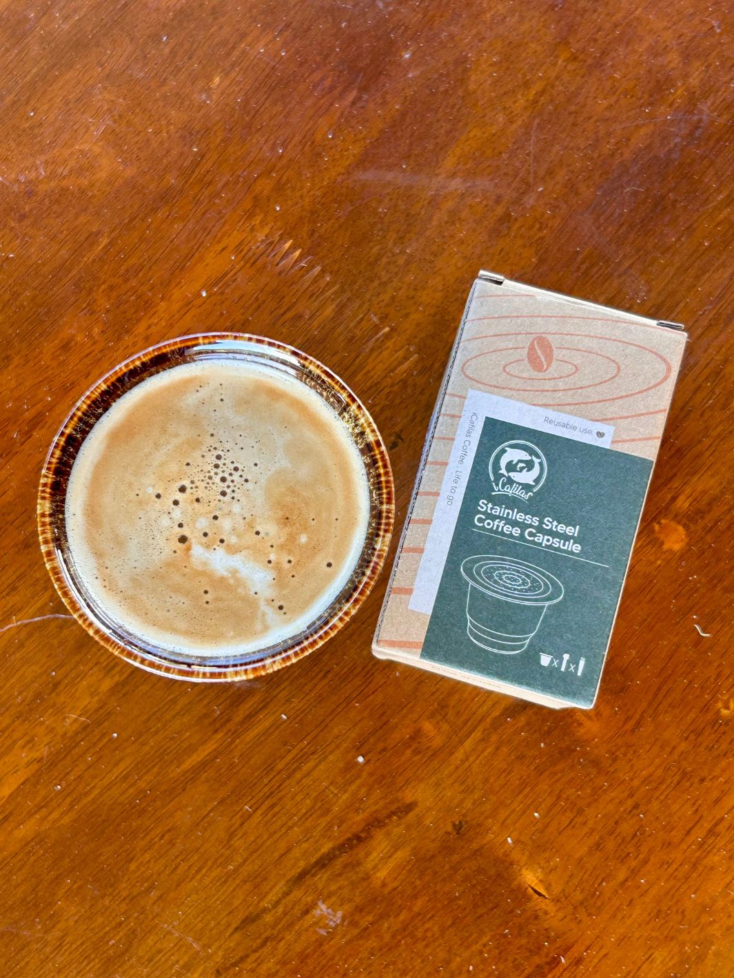 Stainless steel reusable nespresso coffee capsule in packaging next to a cup of coffee on a wooden surface